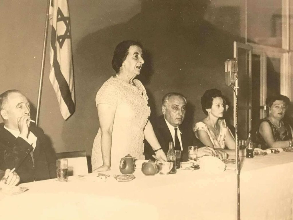 una mesa con personas sentadas reunidas en un vento bnei brithay una mujer parada y al lado tiene la bandera de israel